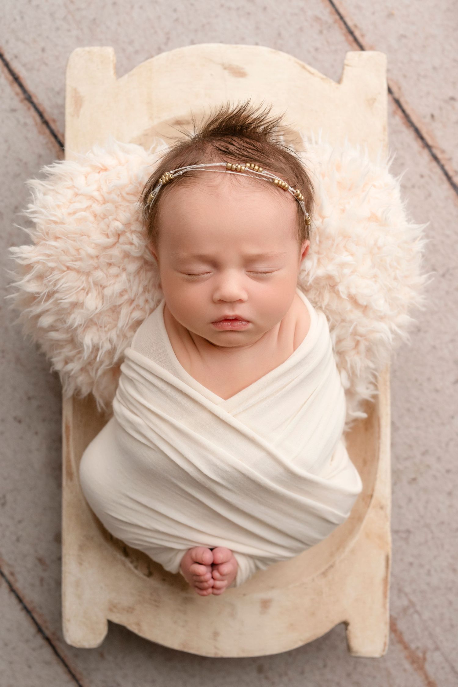 A sleeping newborn swaddled in white fabric lies on a wooden bed with fluffy cream-colored fur padding.