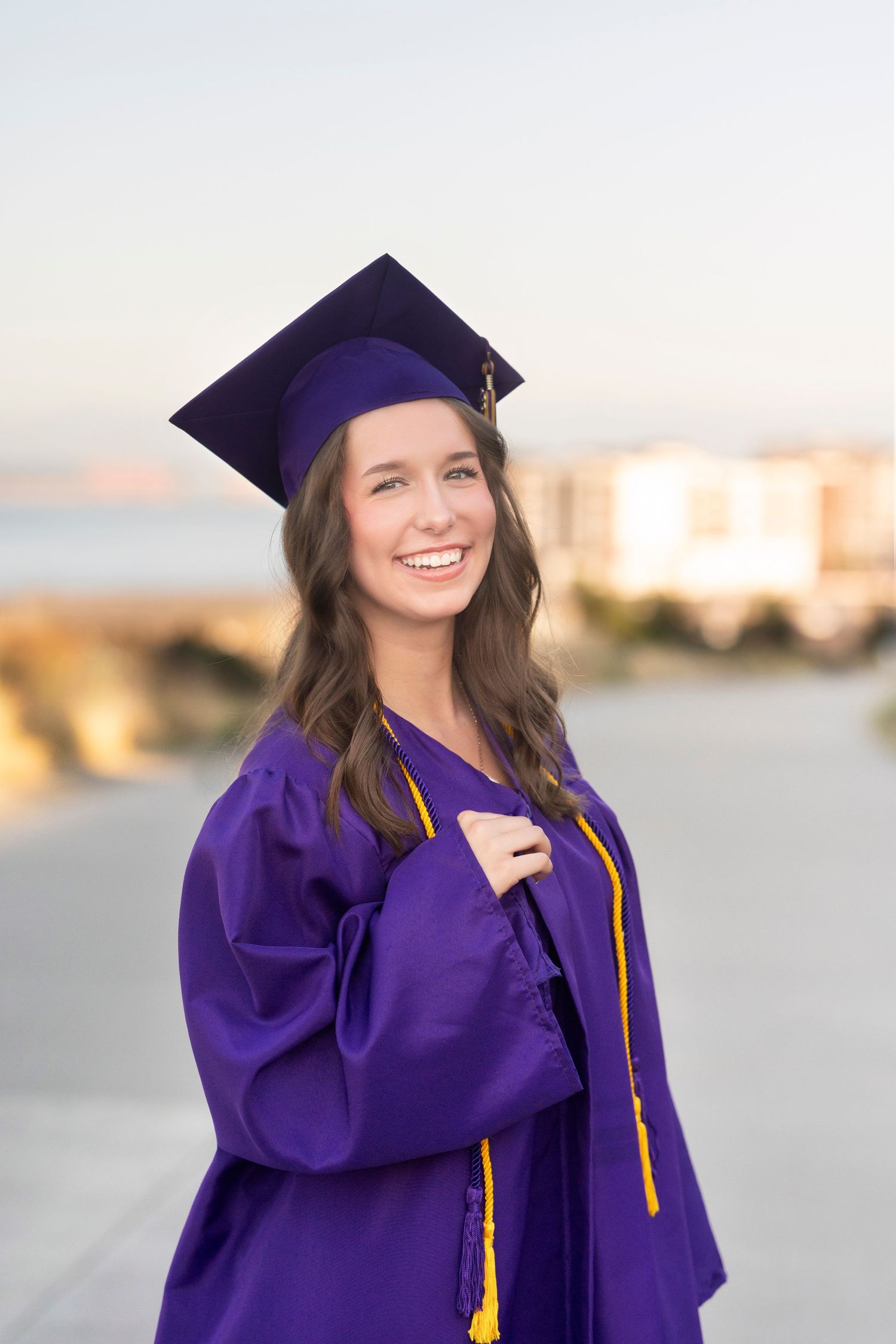Smiling young woman in purple graduation cap and gown posing outdoors on a bright day.