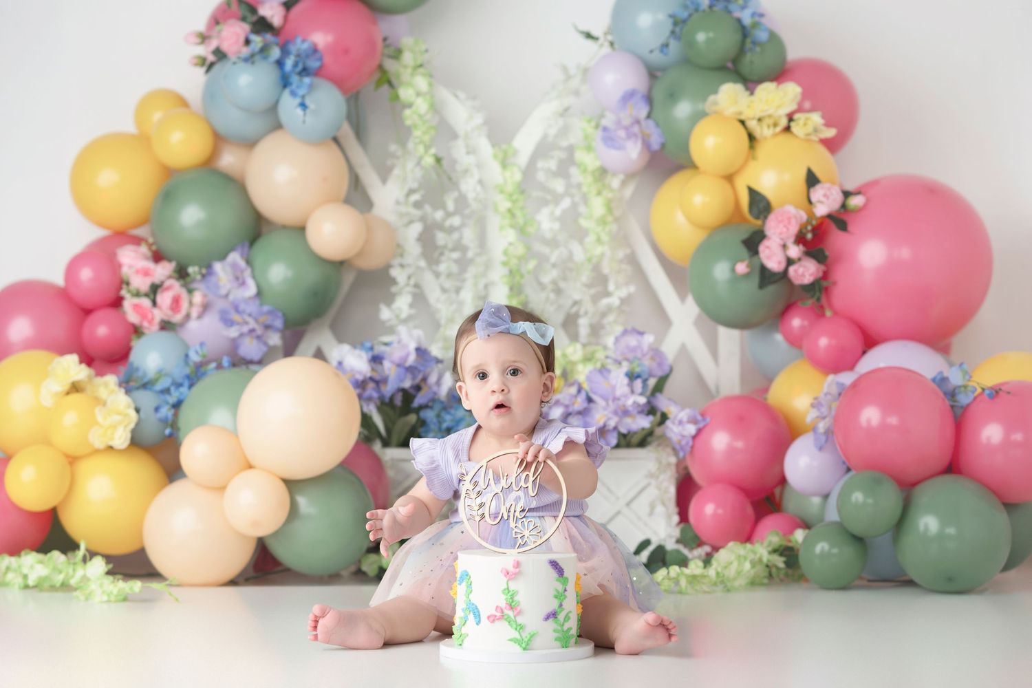 Colorful balloon arch installation with pastel pink, blue, yellow and sage green balloons against a white wall.