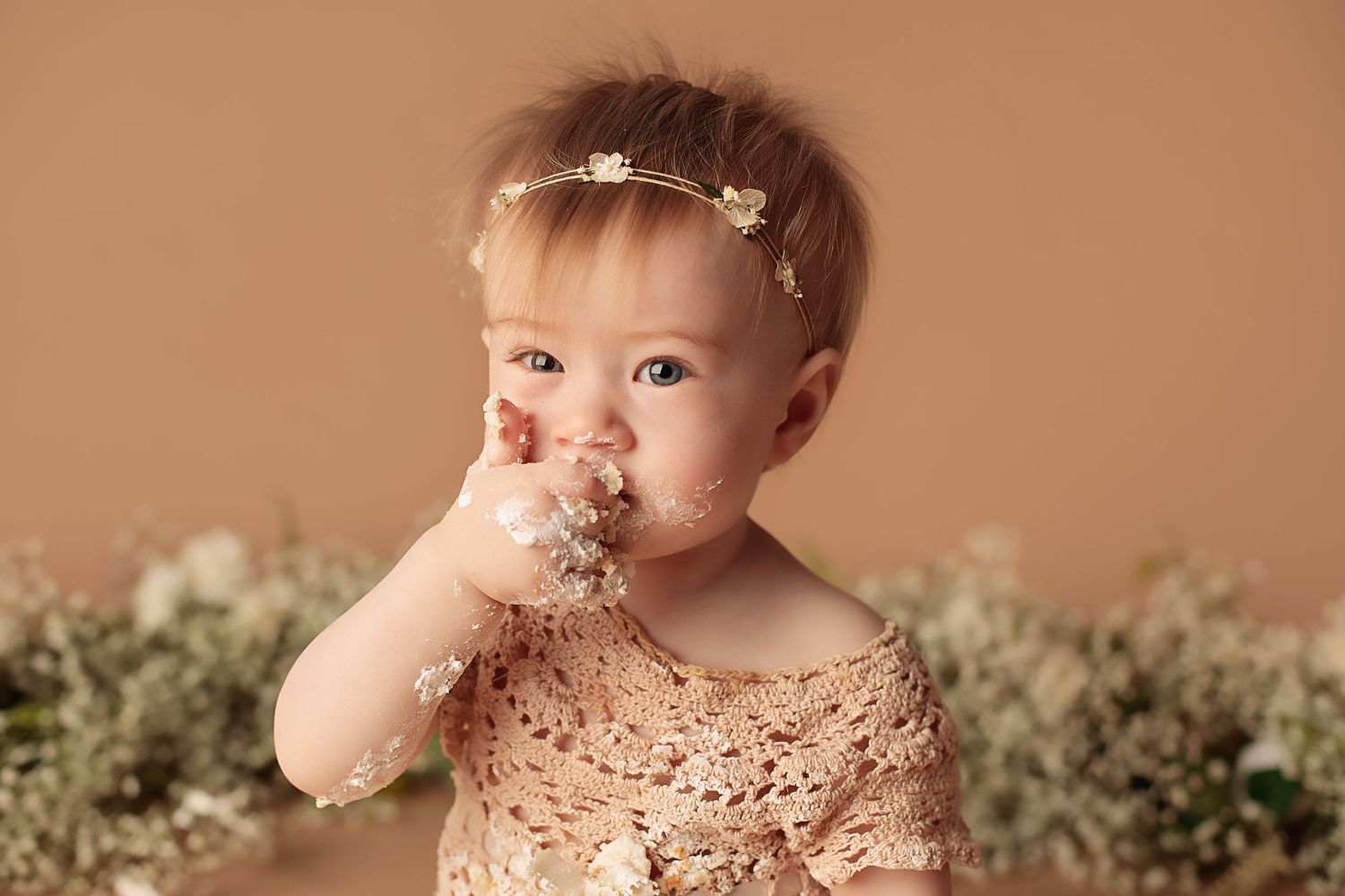 Baby wearing lace dress and delicate headband takes a big bite of cake during cake smash photo session.