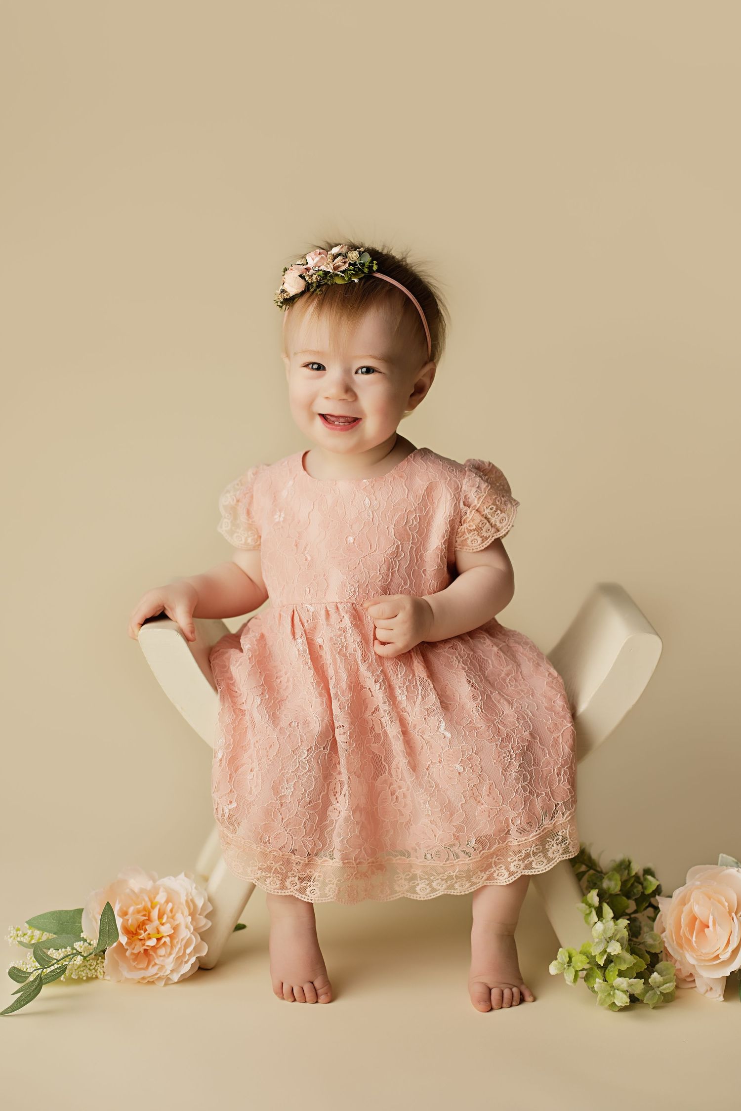 Baby in pink lace dress sits on beige backdrop with peach-colored flowers and greenery for milestone photo session.
