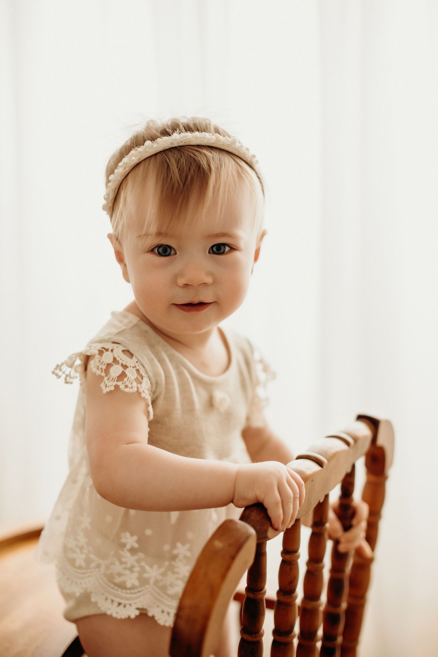 A baby in a lace dress smiles while standing against a white railing in bright natural light.