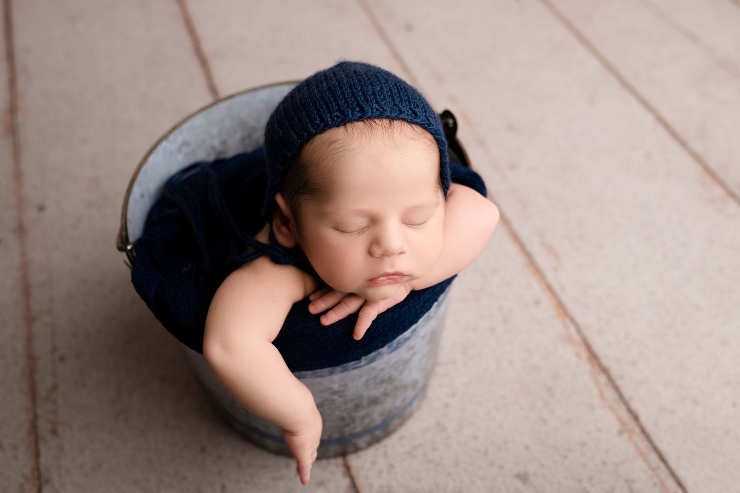 Baby resting in dark blue fabric inside grey bowl on light wooden floor.