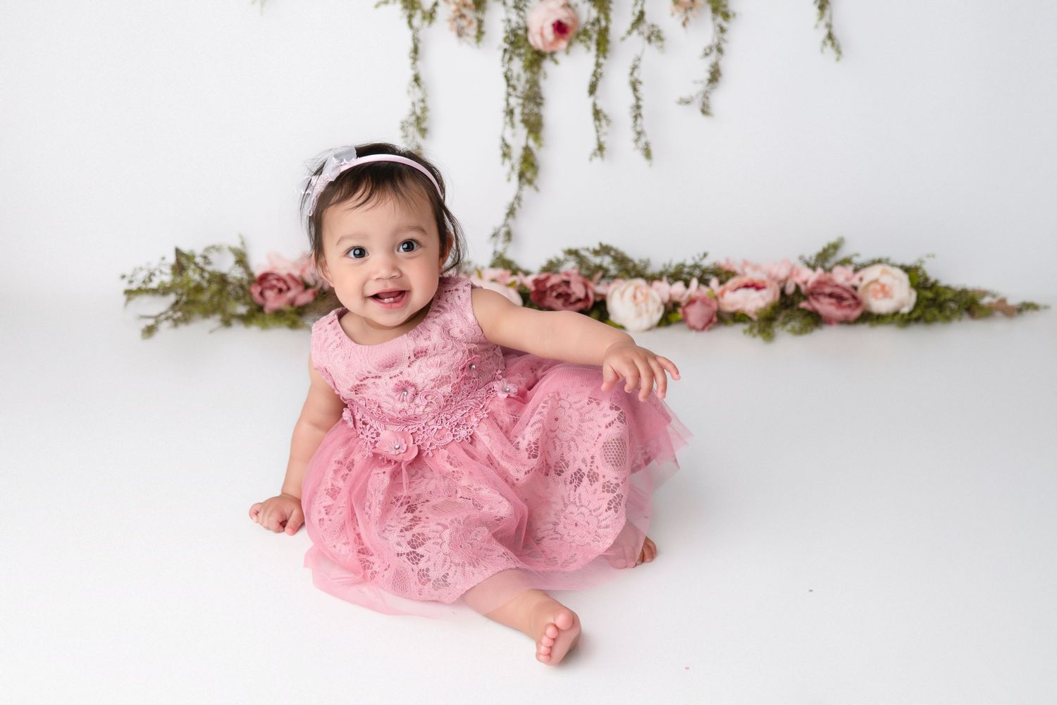 Baby in pink lace dress lies on white floor with floral decorations overhead.