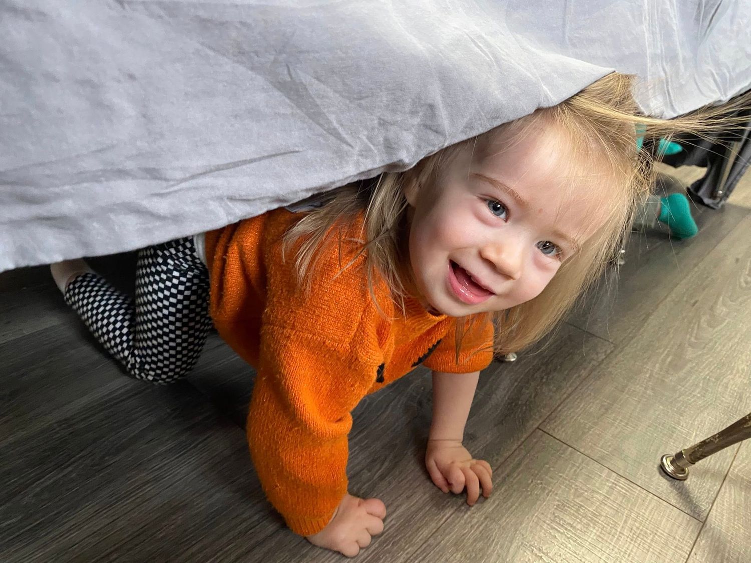 Smiling toddler girl in orange sweater crawling under a bed on hardwood floors.