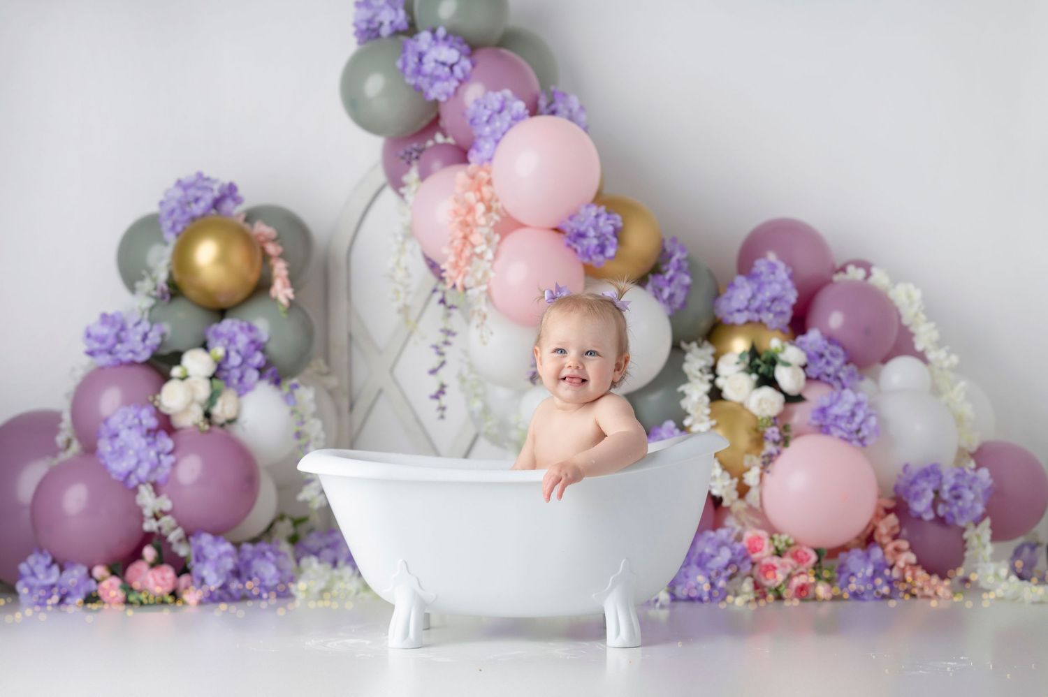 Baby smiling in white bathtub surrounded by purple flowers and colorful balloons in a festive photo shoot.