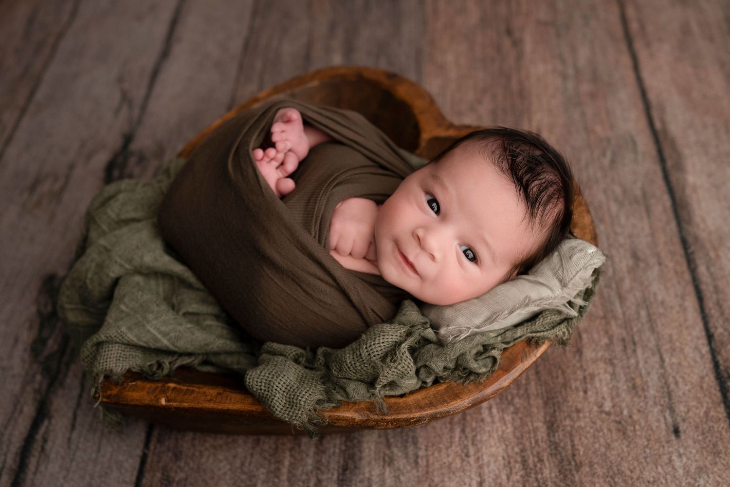 Newborn baby wrapped in olive green swaddle, smiling in a heart-shaped wooden bowl on rustic wood floor.