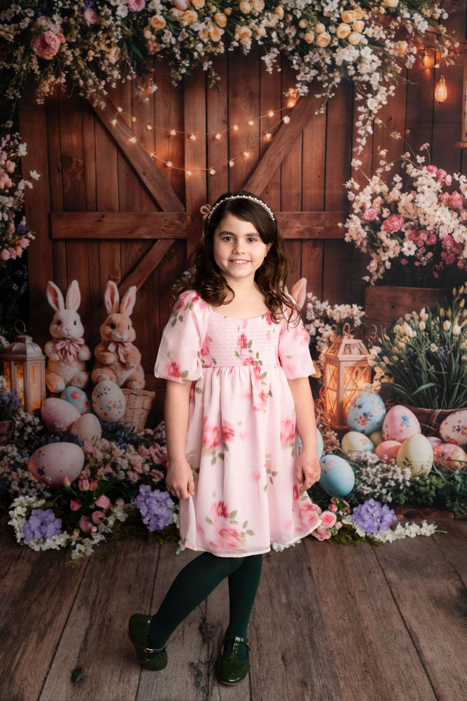 Young girl in pink floral dress poses before Easter backdrop with bunnies, colorful eggs, flowers, and barn doors.
