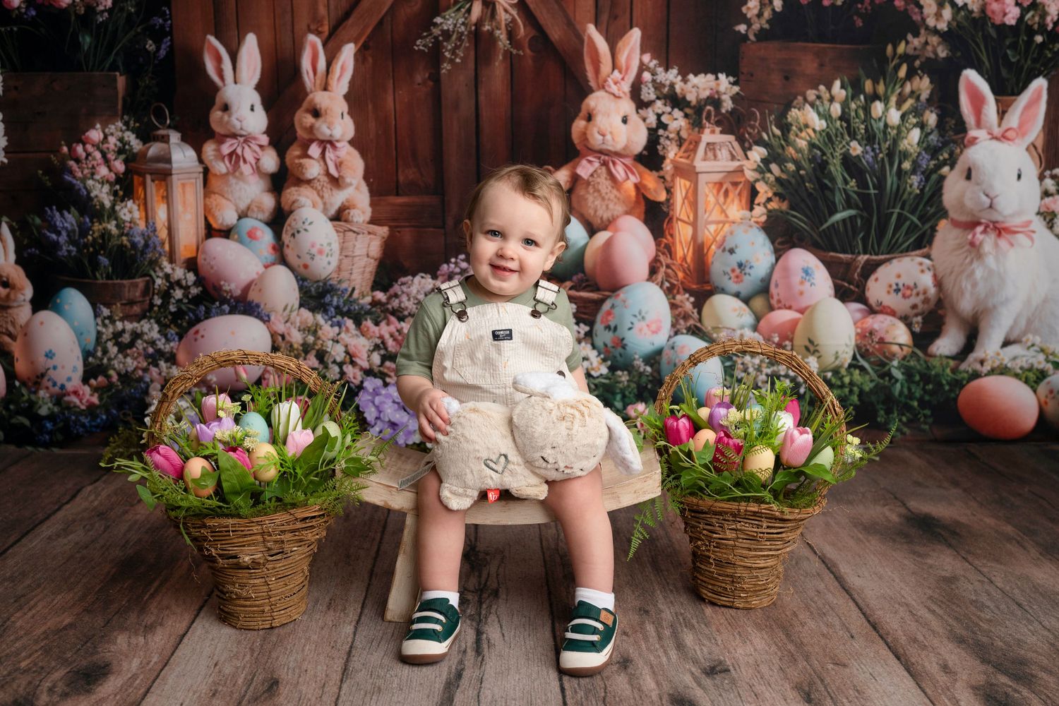 Toddler in overalls holding a stuffed bunny surrounded by Easter eggs, baskets, and decorative rabbits on a wooden backdrop.