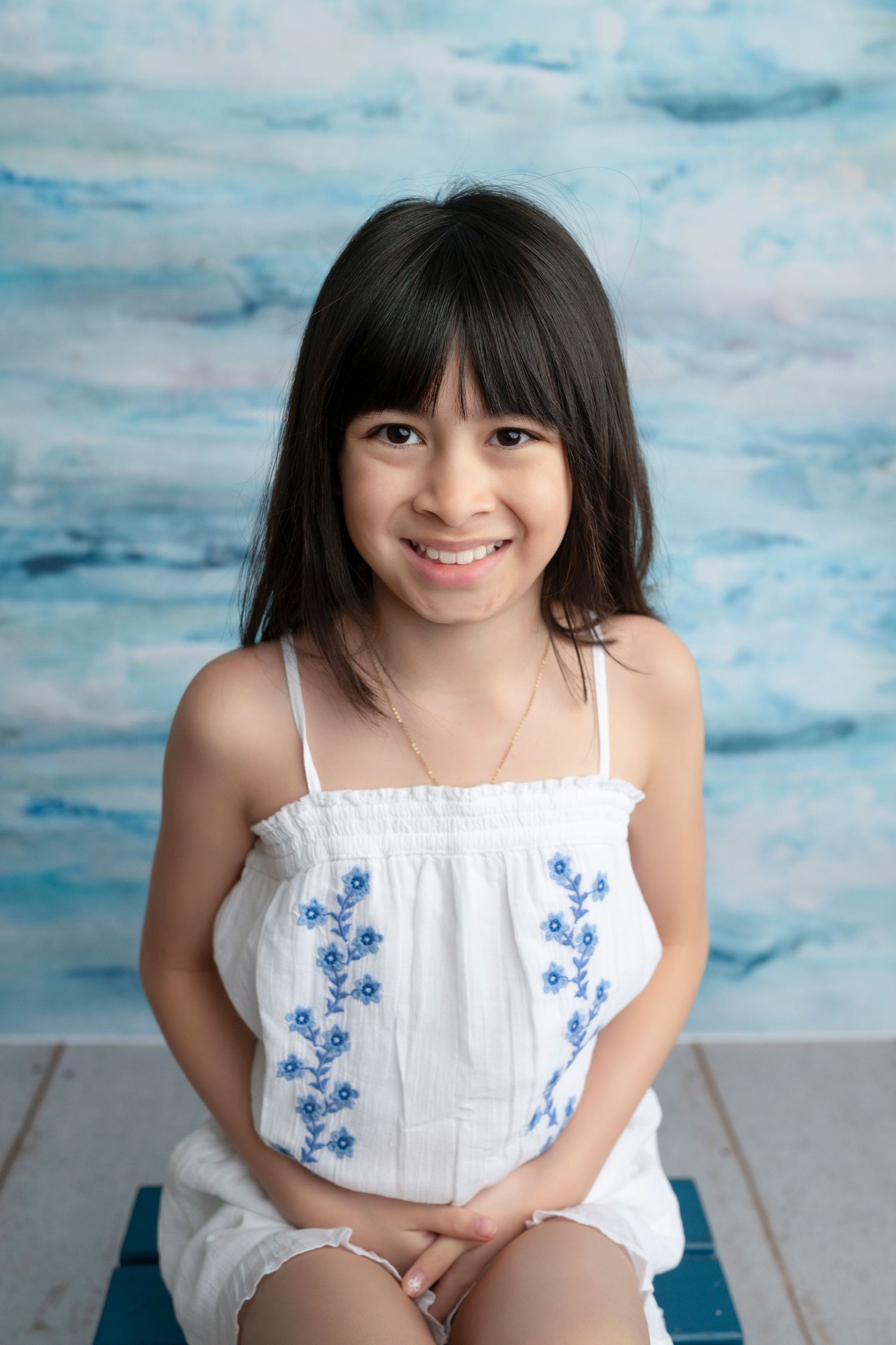 Smiling young girl with dark hair wearing a white sundress with blue floral embroidery, posed against a blue watercolor backdrop.