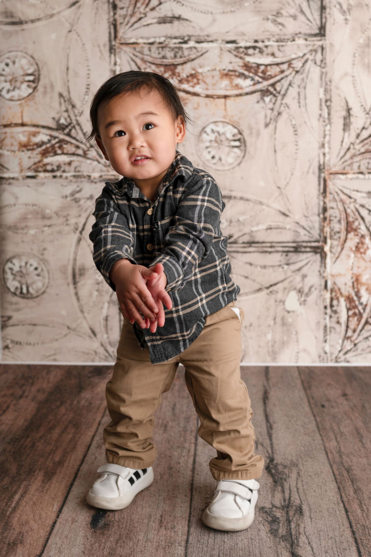 Toddler boy in plaid shirt and khaki pants poses in front of vintage decorative backdrop on wood floor.