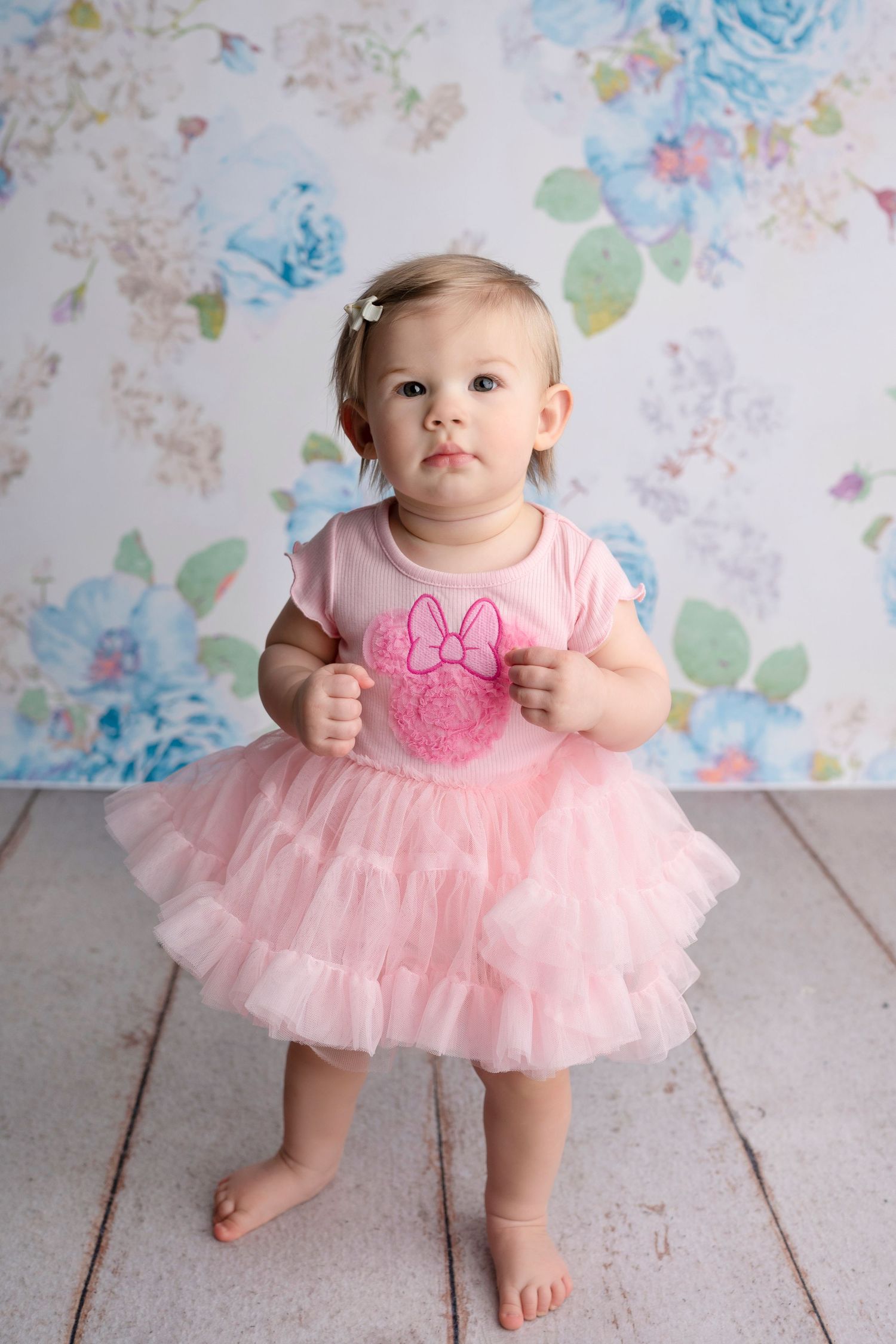 Adorable baby girl in pink tutu dress with Minnie Mouse print stands barefoot against floral backdrop.
