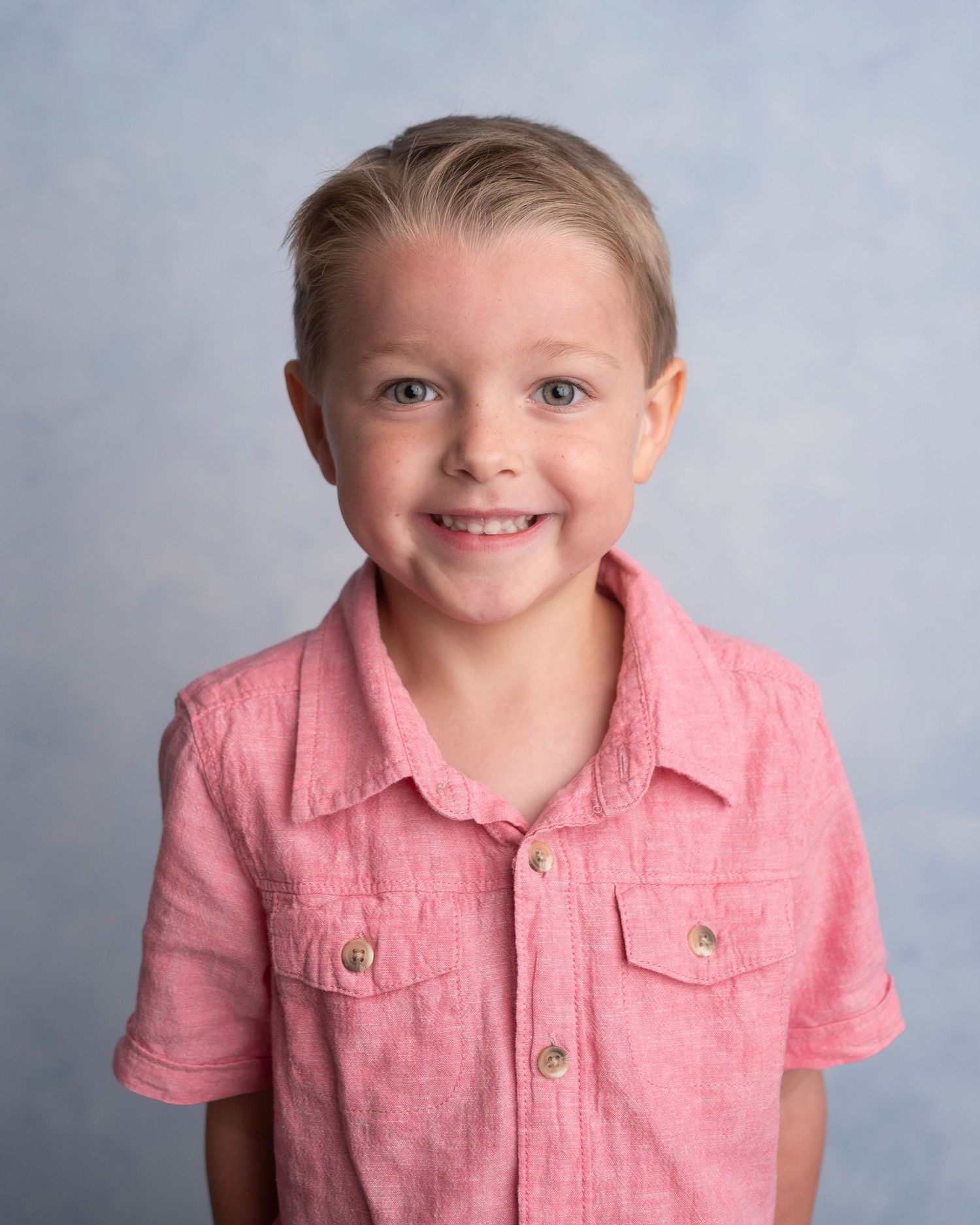 Smiling young boy with blonde hair wearing a pink button-up shirt, posing for a school portrait photo.