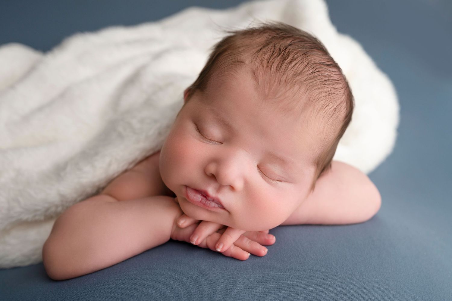 Peaceful newborn baby sleeping on blue surface, hands tucked under chin, wrapped in soft white blanket.