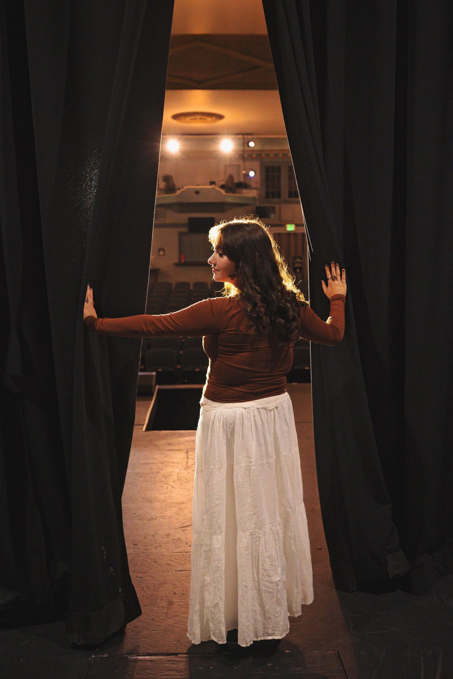 A figure in a white flowing skirt and brown top performs an expressive dance movement between dark curtains.
