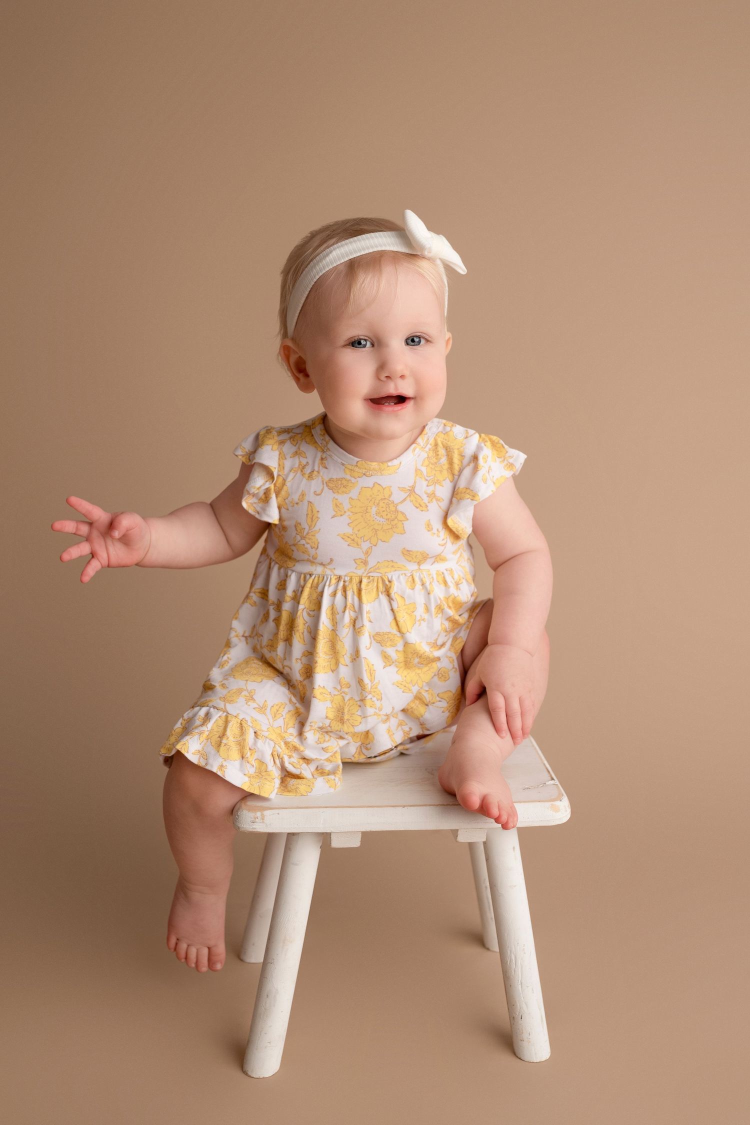 Baby in yellow floral dress sitting on small white stool in studio setting with brown background.