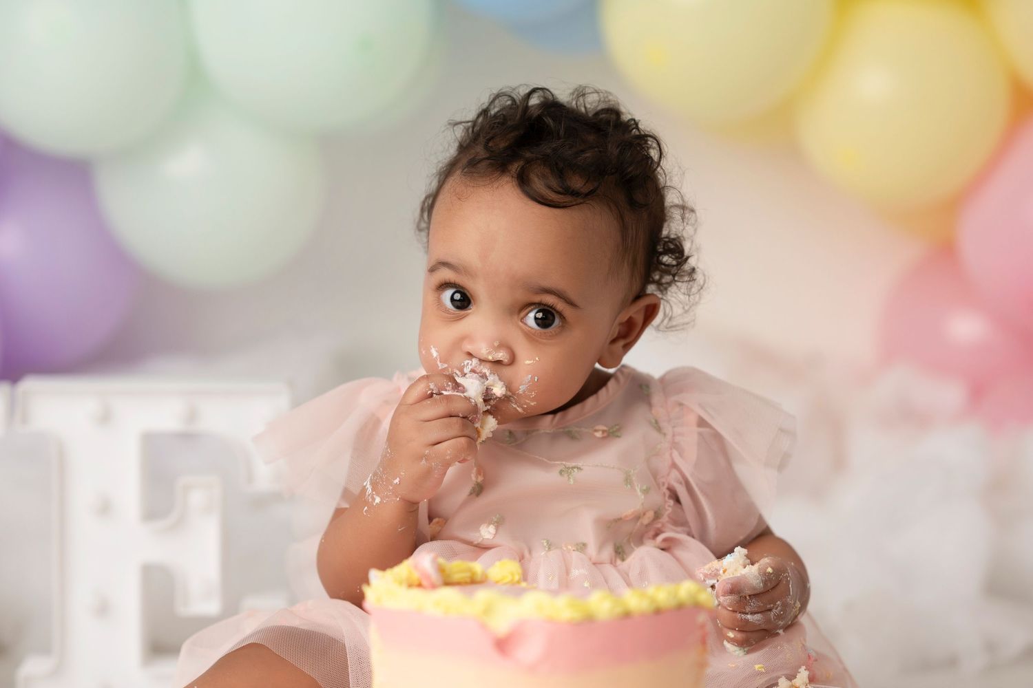 A baby in a pink dress celebrates their first birthday with a smash cake against a pastel balloon backdrop.