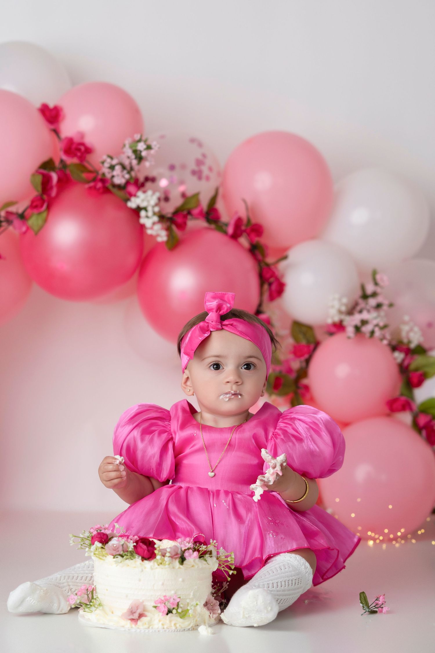 Pink and white balloons create a dreamy backdrop with cherry blossoms and twinkling lights for a baby photoshoot.