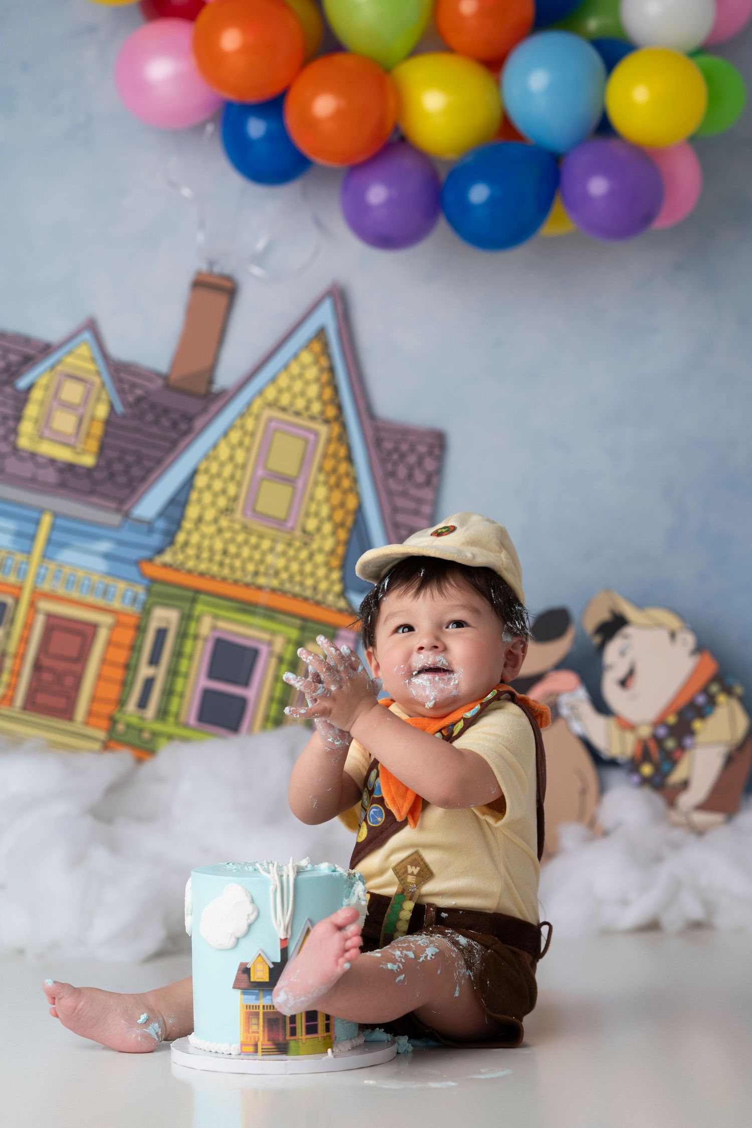 Colorful balloon backdrop with cartoon houses and clouds creates whimsical setting for cake smash photography session.