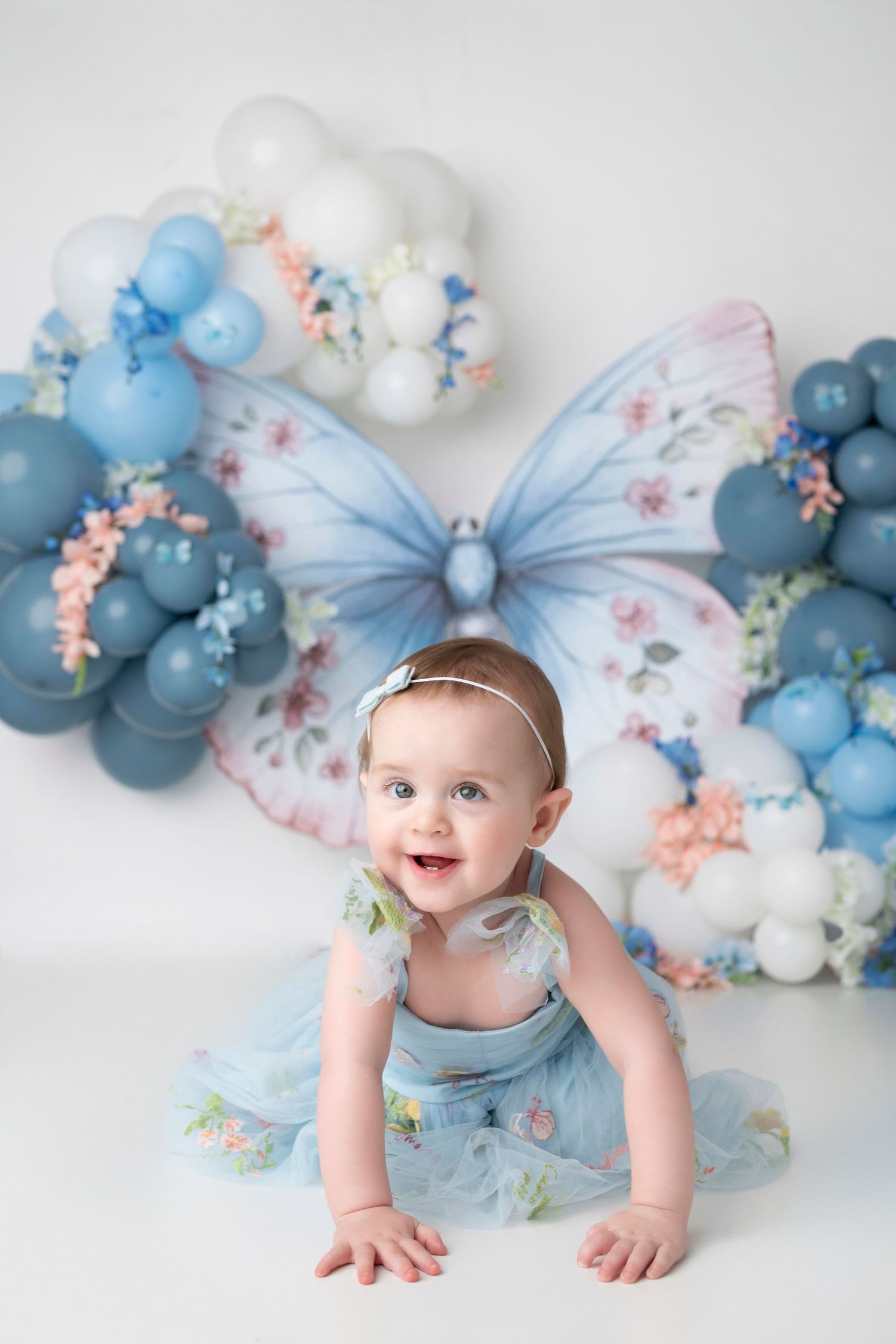 Baby in light blue dress crawls on white backdrop with pastel balloon arch decoration.