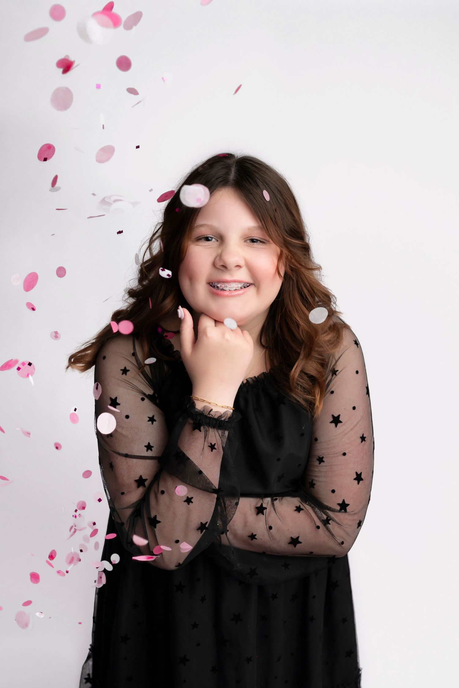 A smiling young girl in a black star-patterned dress poses with falling pink confetti against a white background.