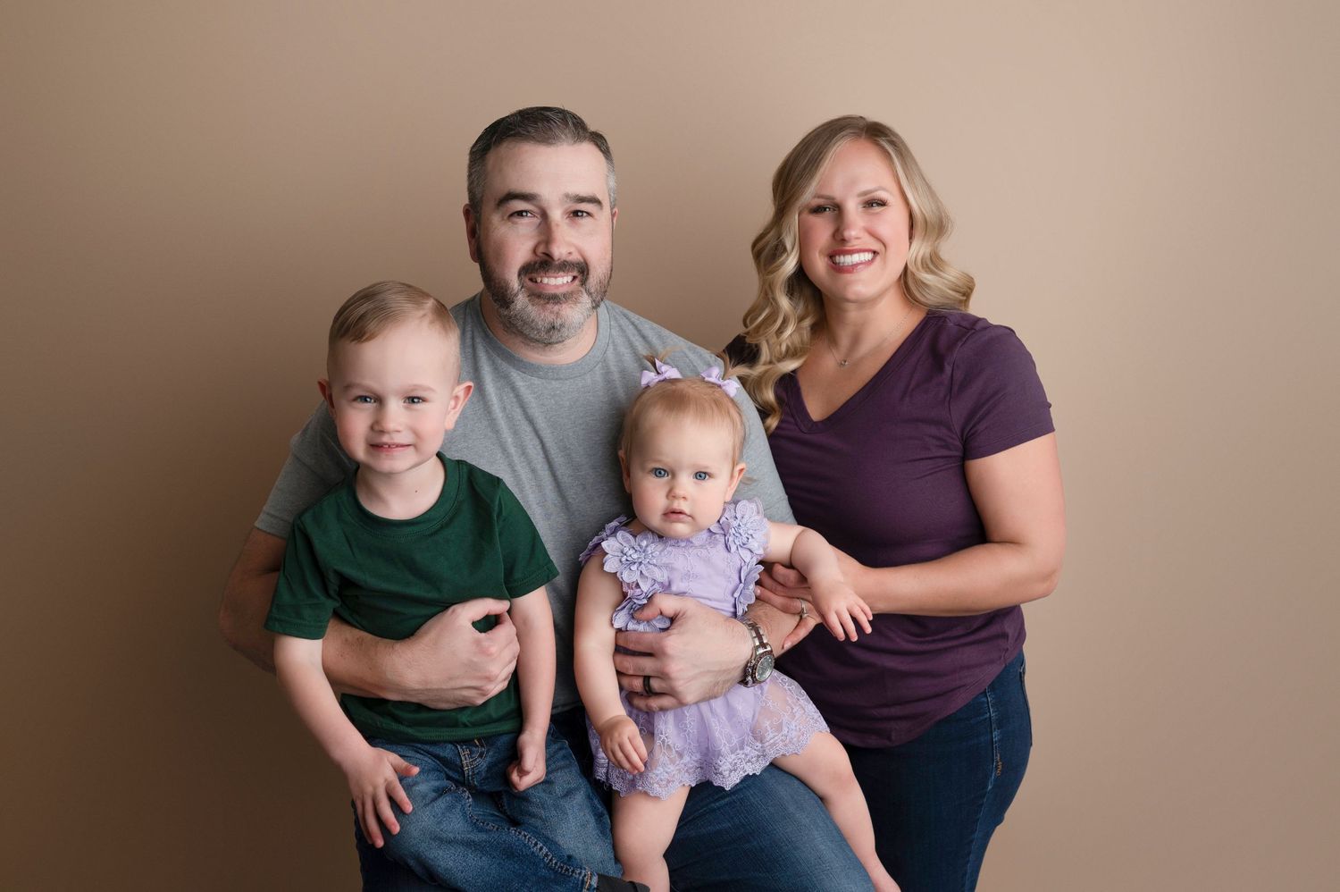 Family of four posing for portrait, parents holding toddler boy and baby girl in purple dress on beige background.