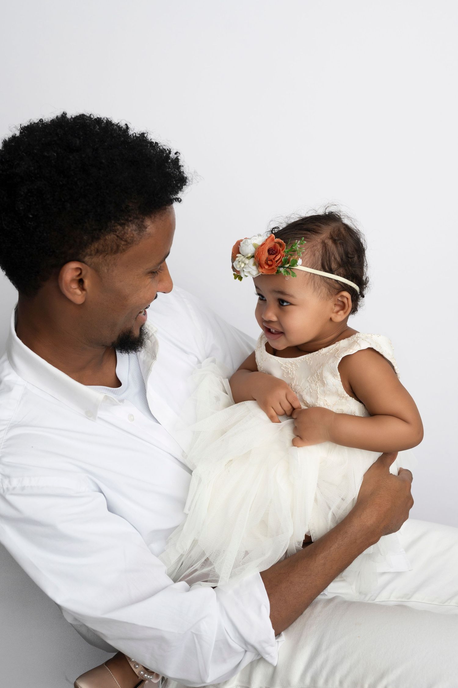 A father in white holds his baby girl in a white tutu dress and floral headband, sharing a joyful moment.