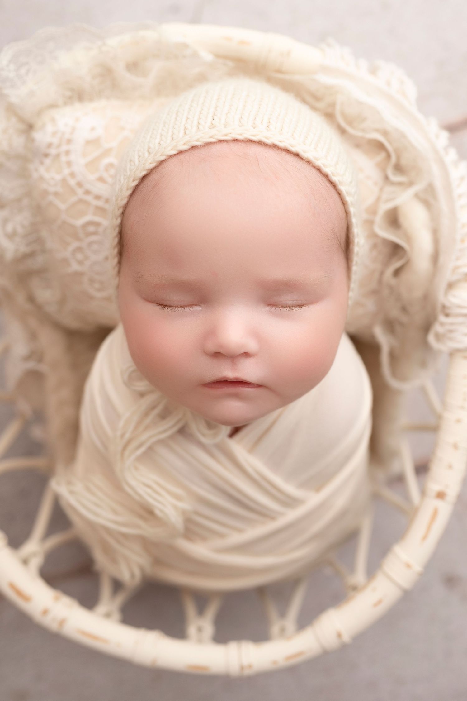 Newborn baby swaddled in cream wrap wearing knit bonnet, sleeping peacefully in white wicker basket.