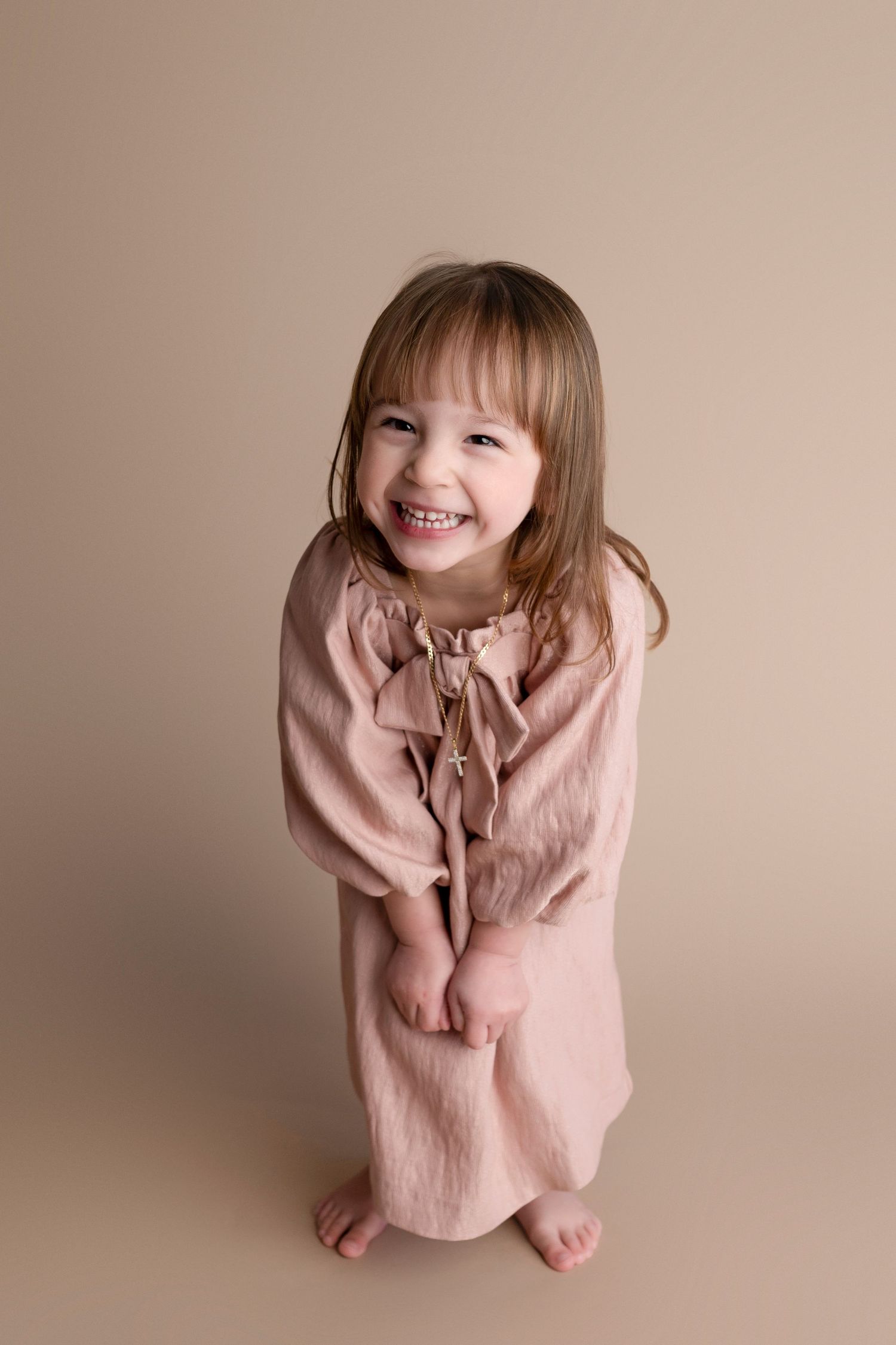 Smiling young girl in pink dress poses barefoot against beige background in a professional portrait photo.