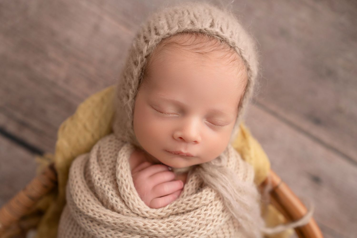 Sleeping newborn baby wrapped in a cozy knit blanket and bonnet, nestled in a basket on a wooden background.