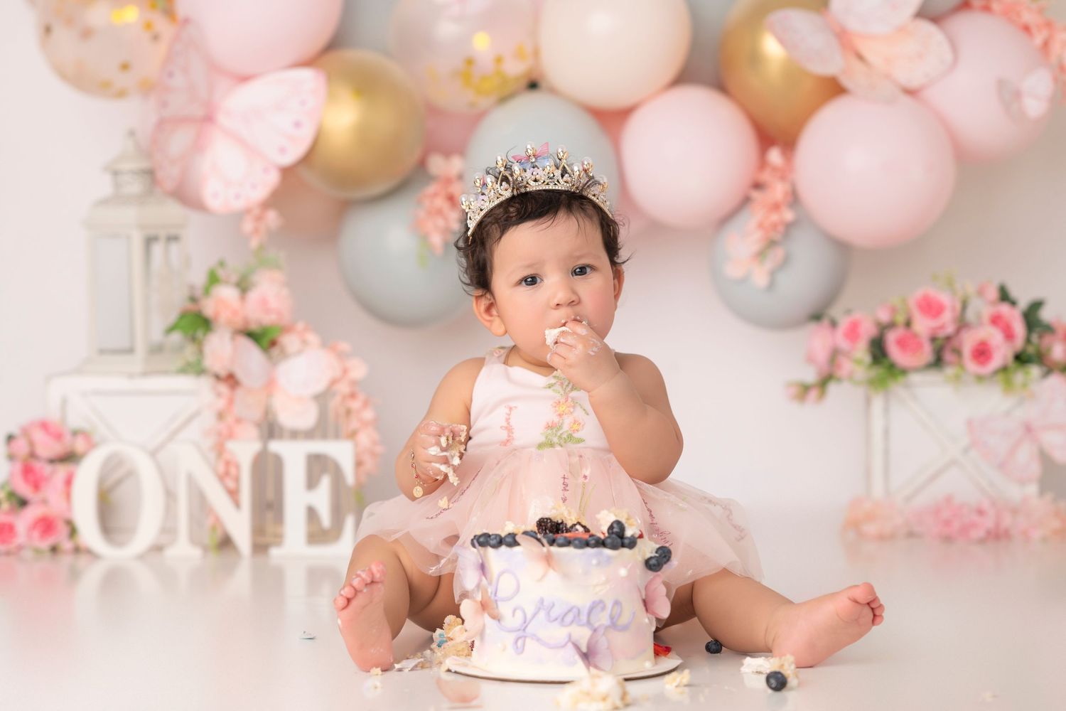 Baby girl in pink dress with crown celebrates first birthday smashing cake surrounded by balloons and floral decorations.