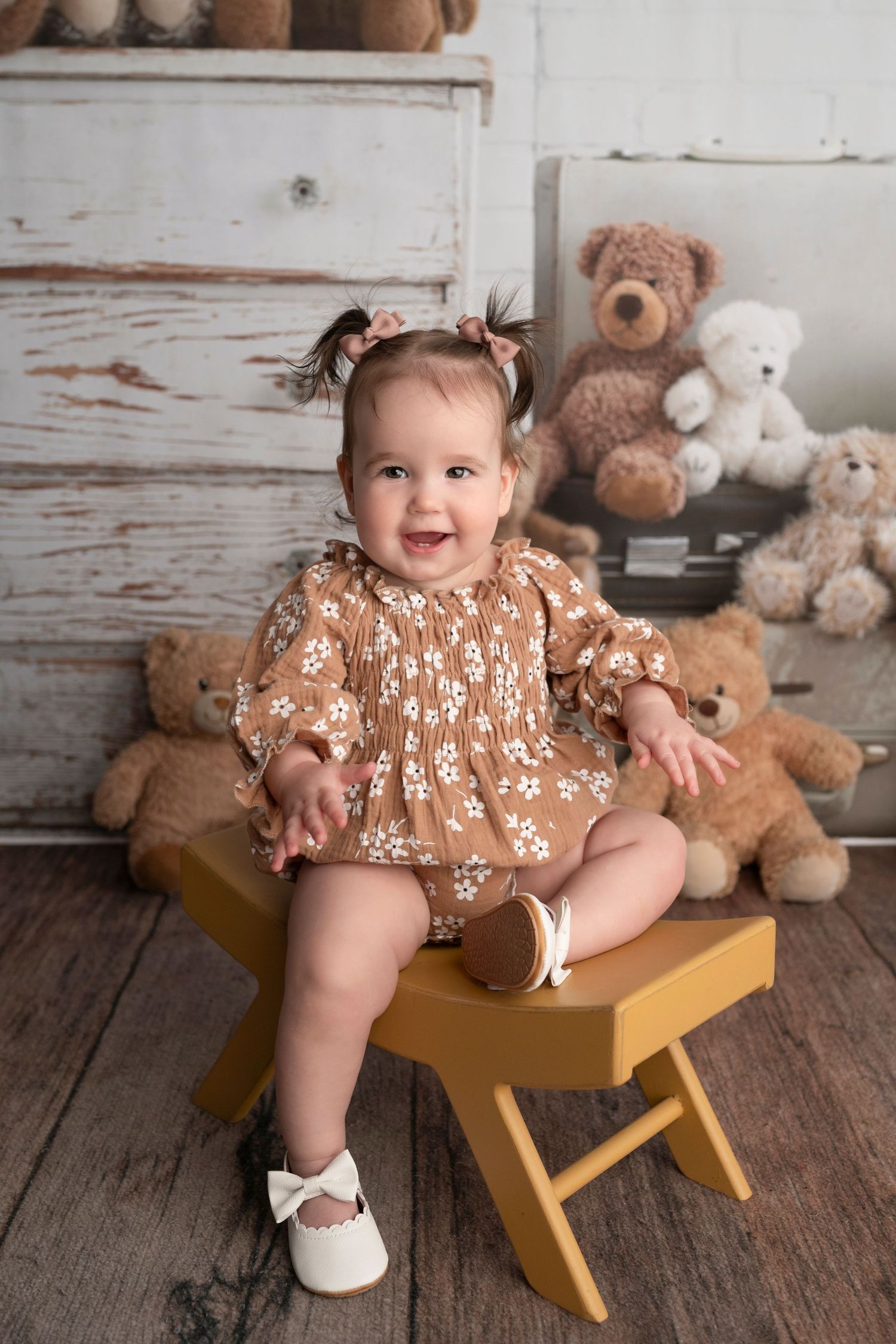 Smiling baby girl in brown floral dress sits on yellow stool surrounded by teddy bears in a rustic photo studio.