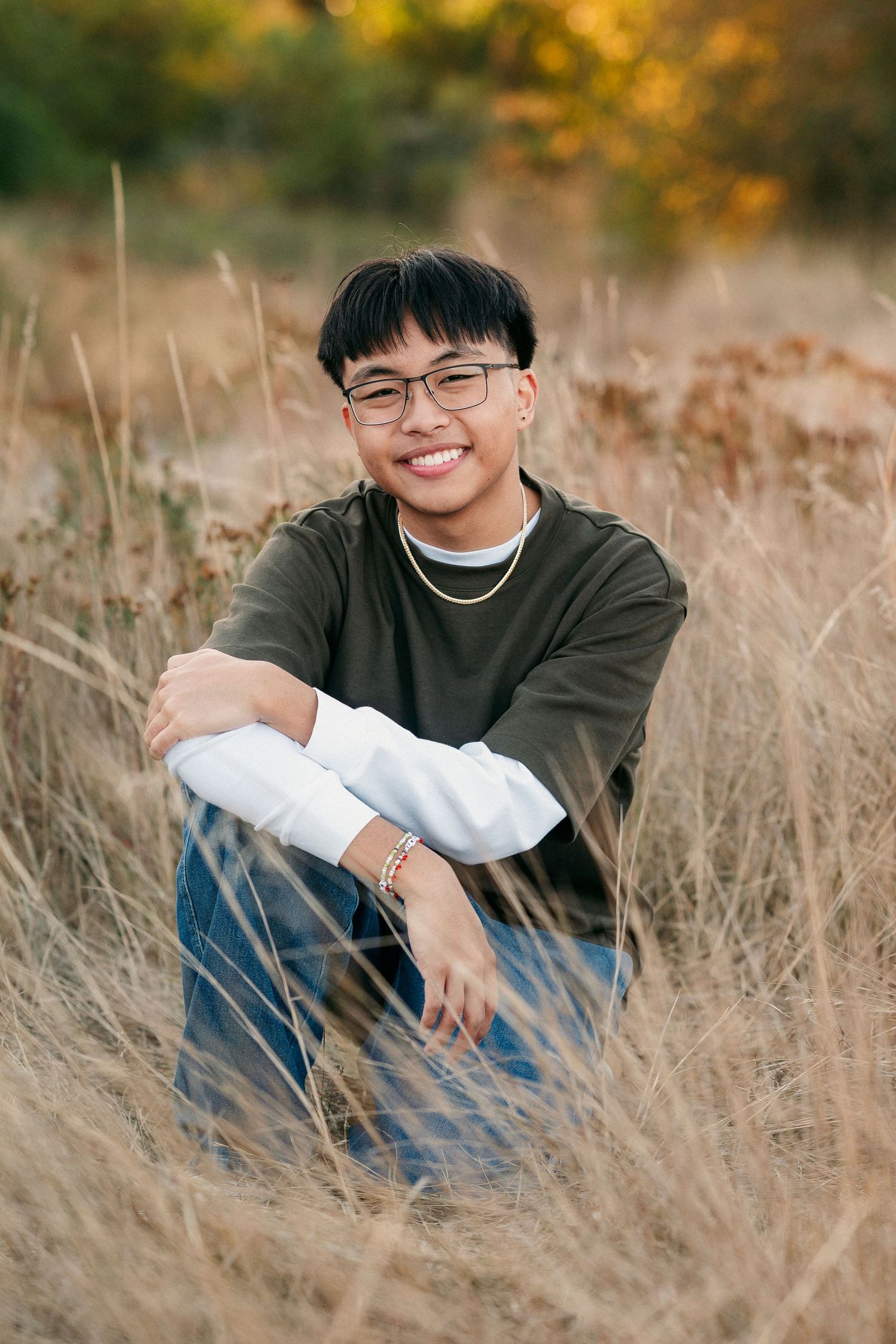 A person wearing glasses and a dark sweater sits among tall golden grass in an autumn field.