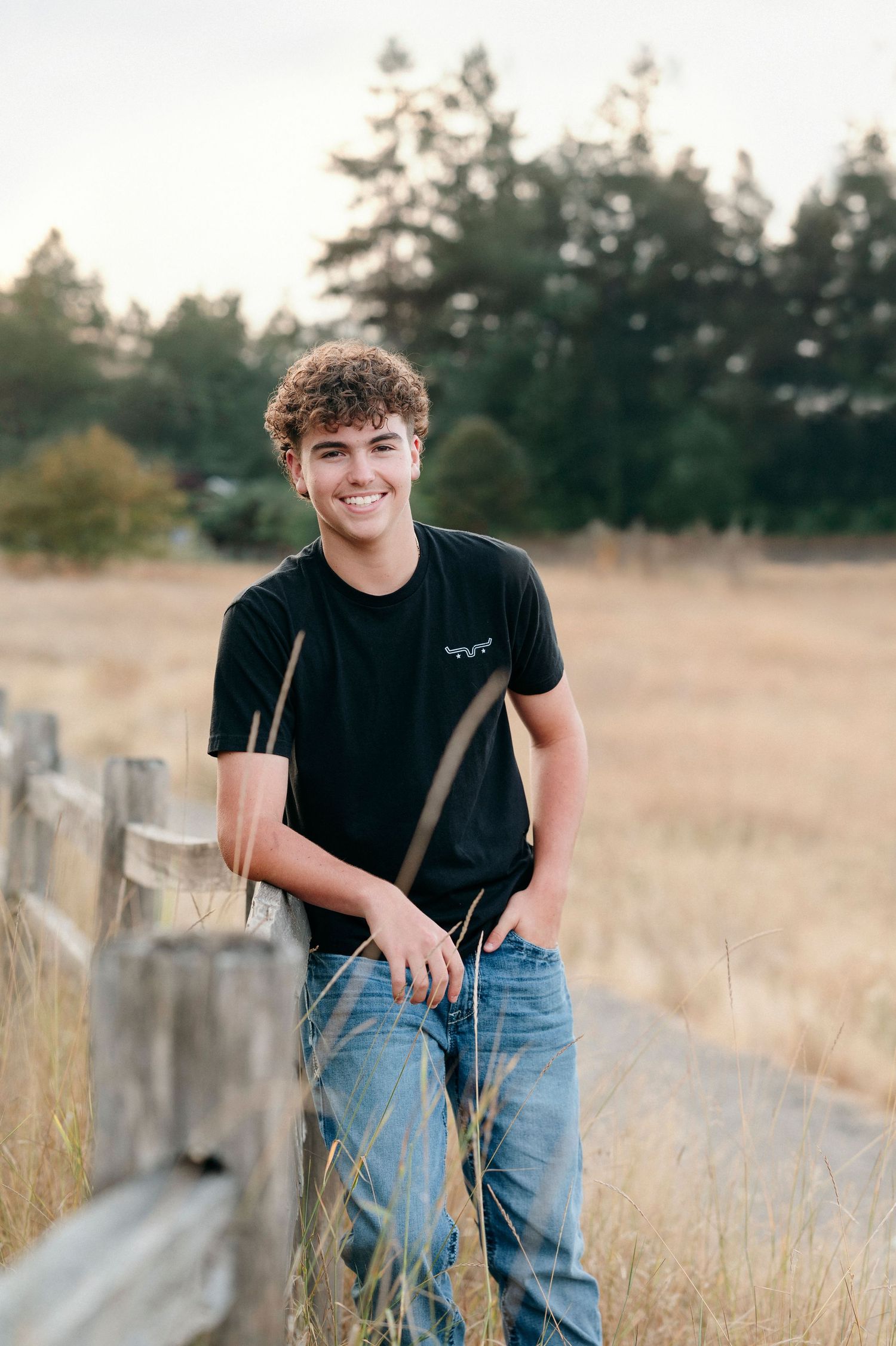 Person in black t-shirt and jeans leaning against rustic wooden fence in golden field at sunset.
