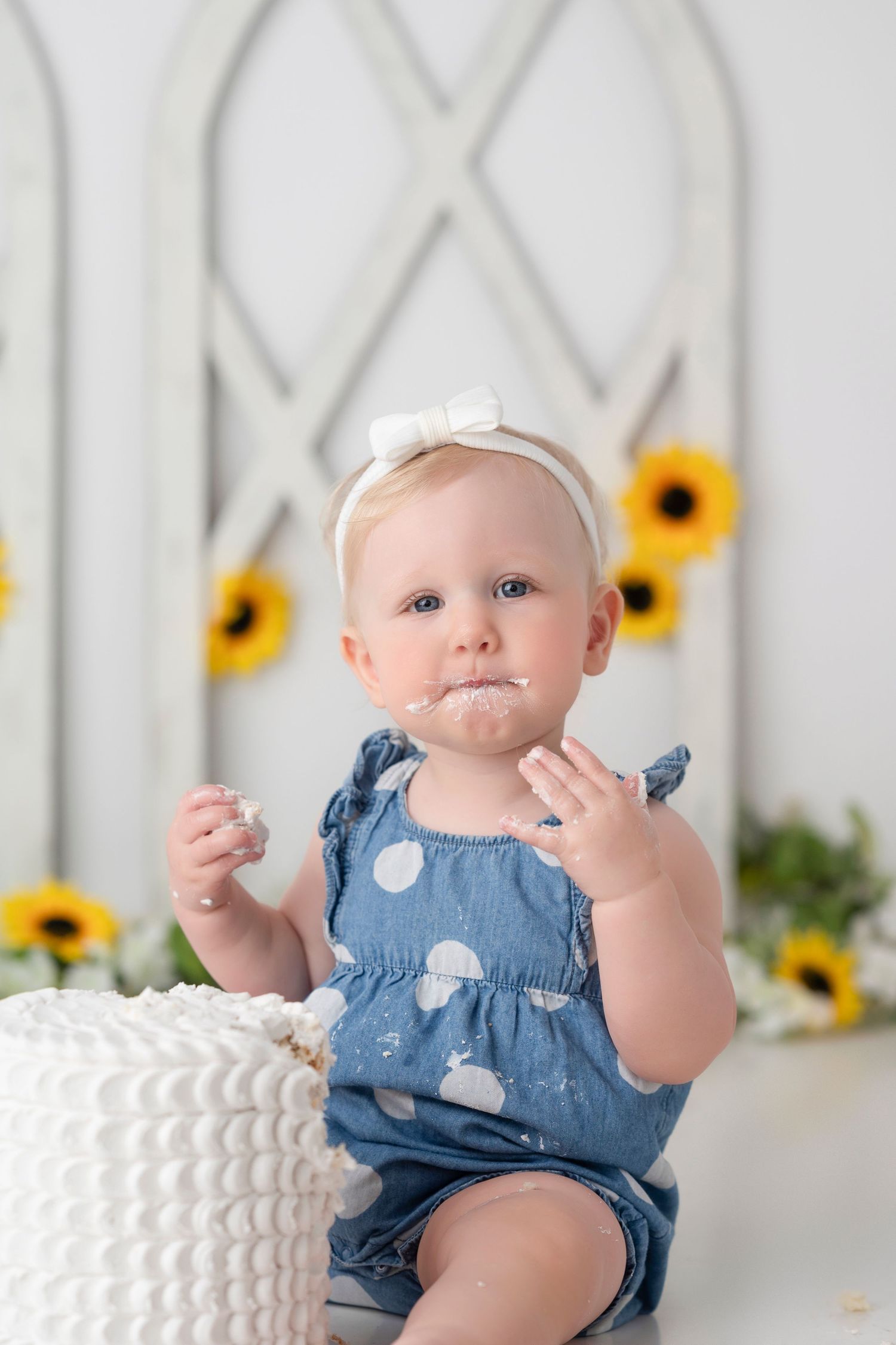 Baby in blue polka dot romper sits next to white cake with sunflower decorations in background.