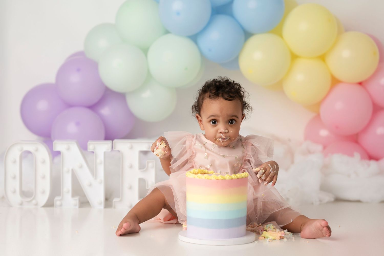 Baby in pastel rainbow cake smash photo session with balloon arch backdrop and white ONE letters.