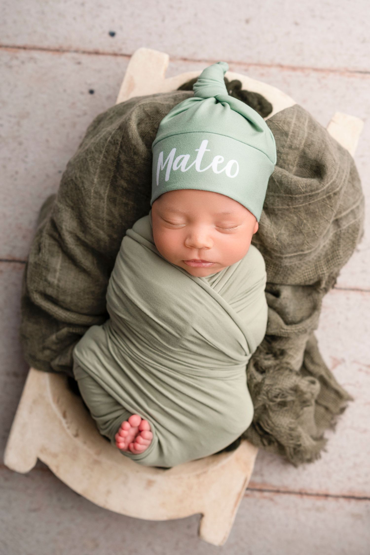 Newborn baby wrapped in sage green swaddle with matching hat sleeping peacefully on textured beige backdrop.