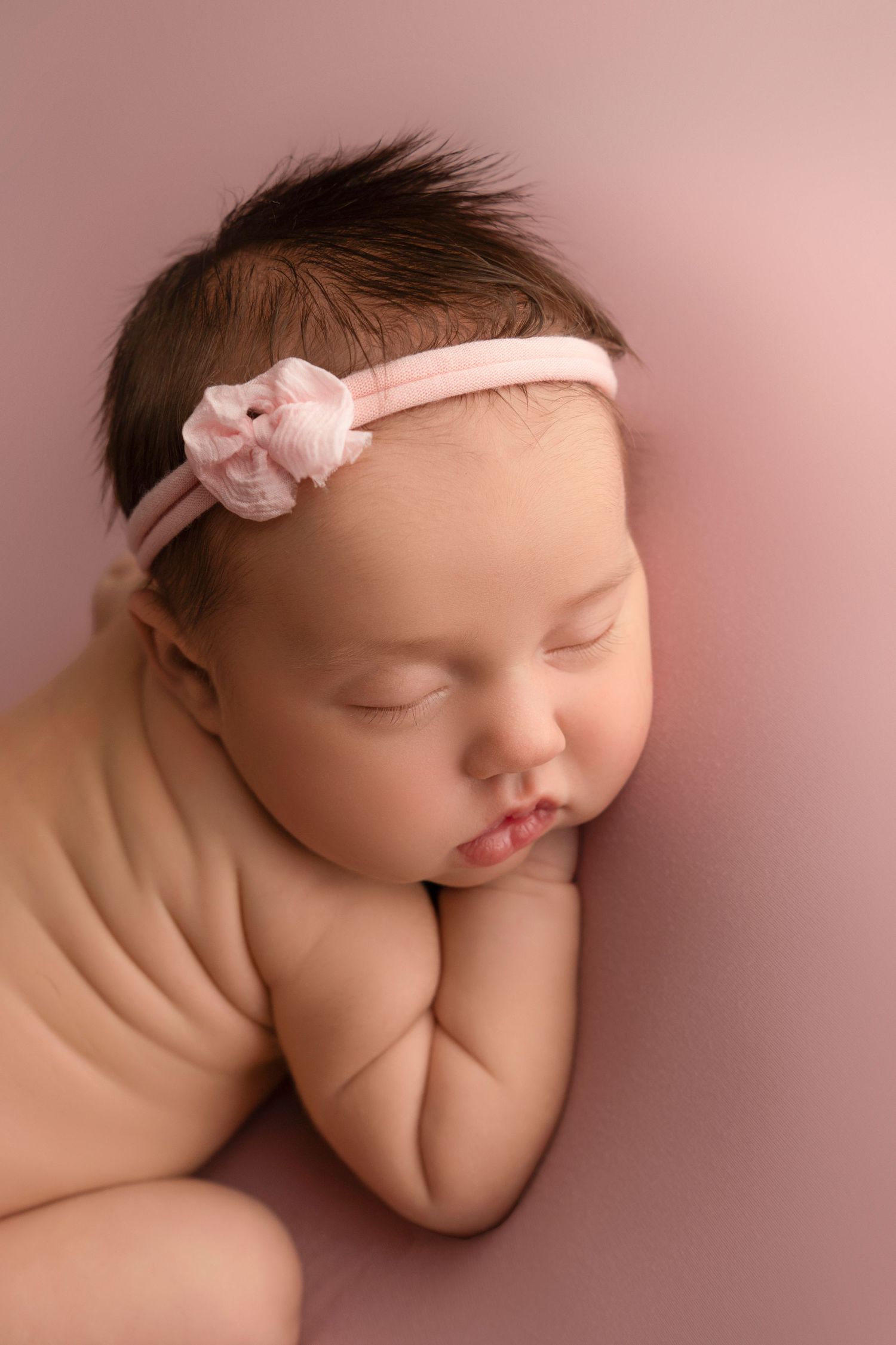 Sleeping newborn in pink headband lying on soft beige blanket in peaceful studio portrait.