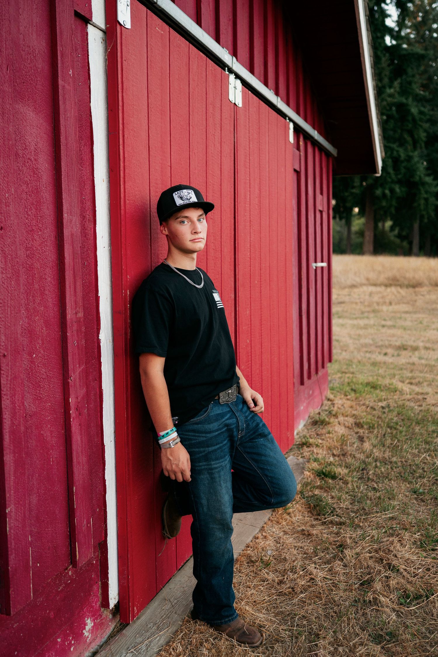A person wearing a baseball cap and black t-shirt leans against a red barn wall in a rural setting.