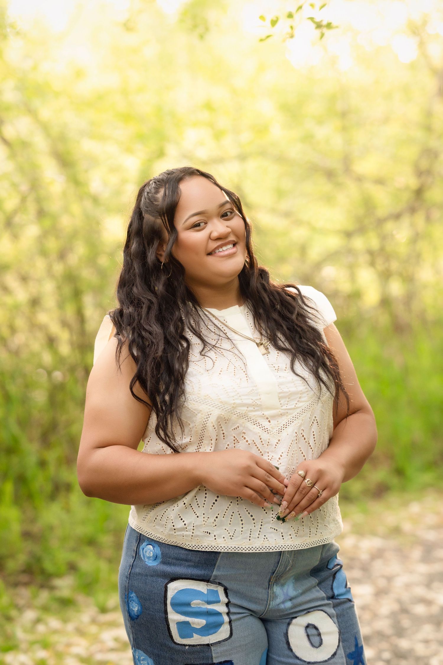 A smiling person wearing a white lace top and distressed jeans poses outdoors against a soft yellow-green background.