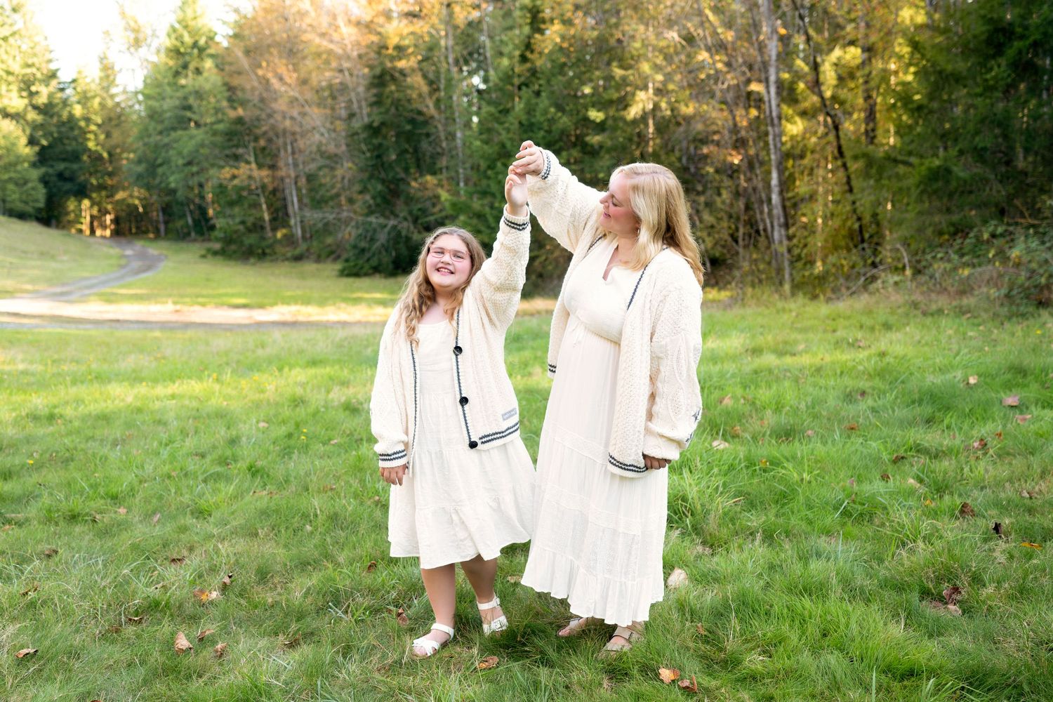 Two people in white dresses walk together through a sunlit grassy meadow at golden hour.