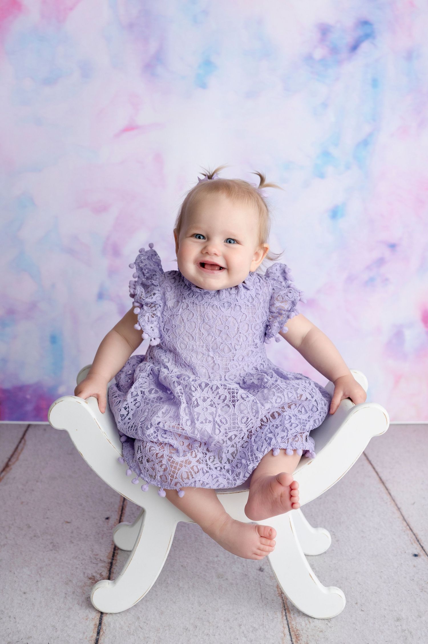 Smiling baby girl in purple lace dress sitting on white chair against pastel watercolor backdrop.