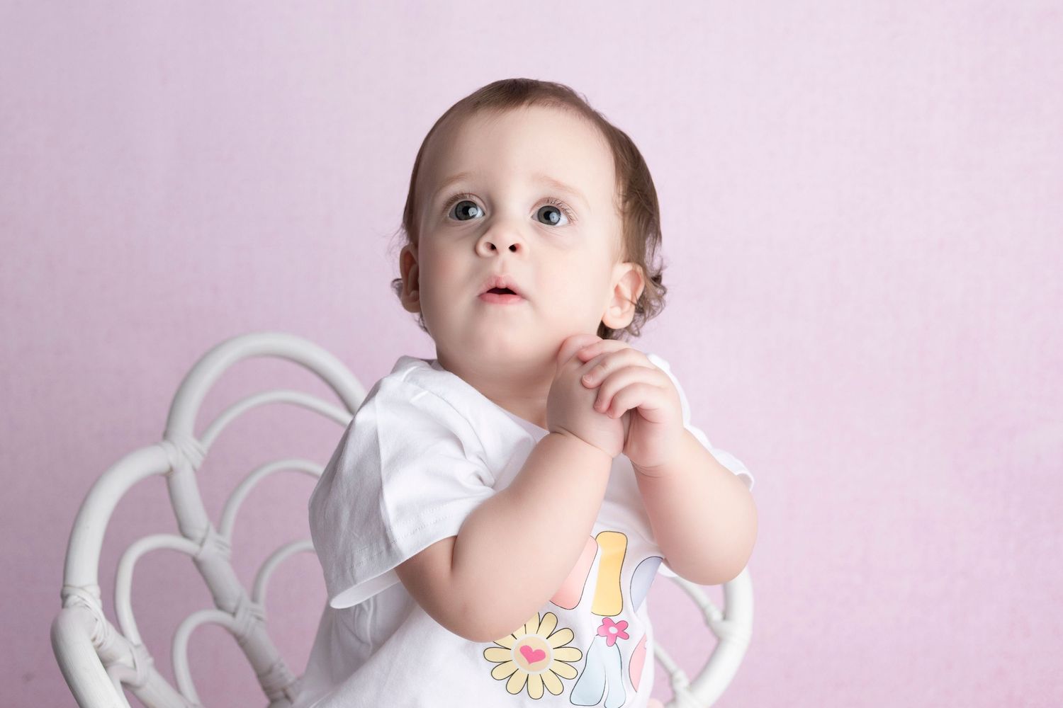 Adorable baby girl with blue eyes sitting in a white wicker chair against a pink background wearing a floral onesie.