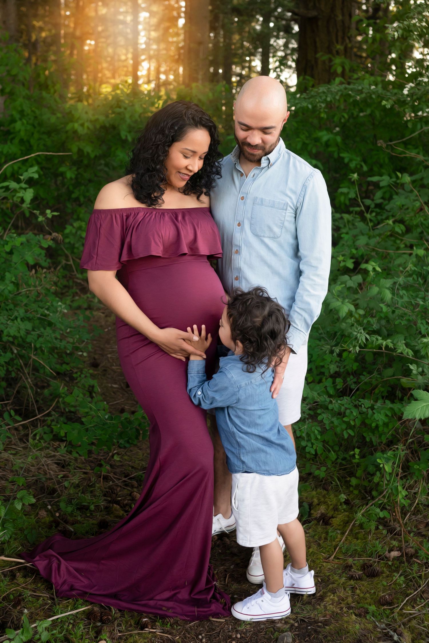 Maternity photoshoot in a forest setting with a burgundy off-shoulder dress and denim shirt at sunset.