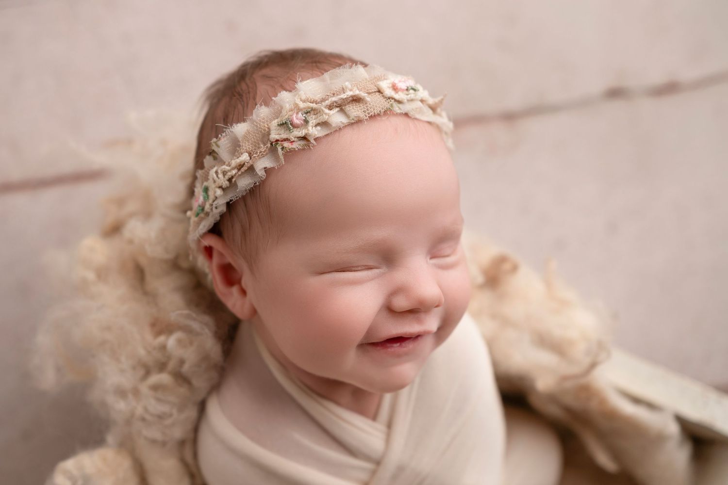 A newborn baby wearing a delicate lace headband smiles while wrapped in soft cream-colored fabric during a photography session.