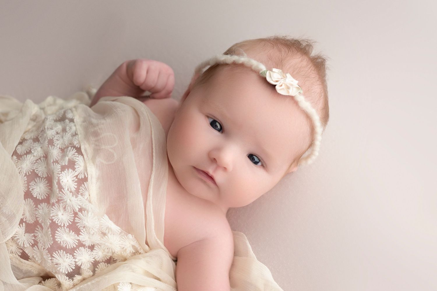 Adorable baby girl in cream lace wrap and floral headband poses for a soft newborn portrait photography session.