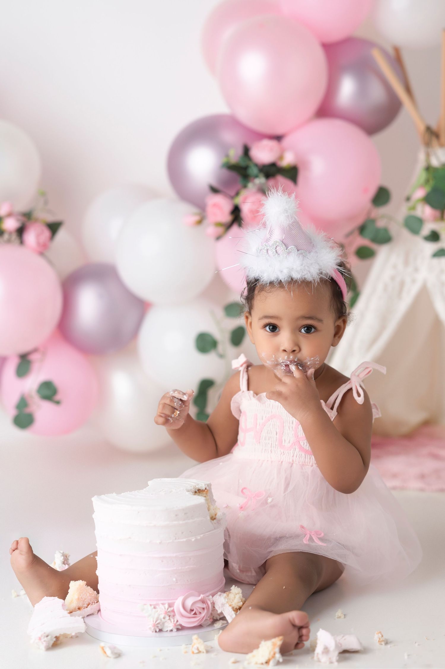 A baby girl in a pink tutu and party hat smashes her first birthday cake surrounded by pink and silver balloons.