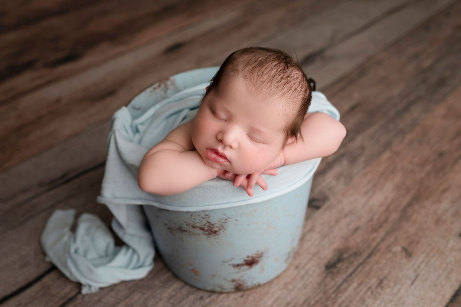 Sleeping newborn baby posed in a rustic blue bucket wrapped in a soft teal cloth on a wooden floor.