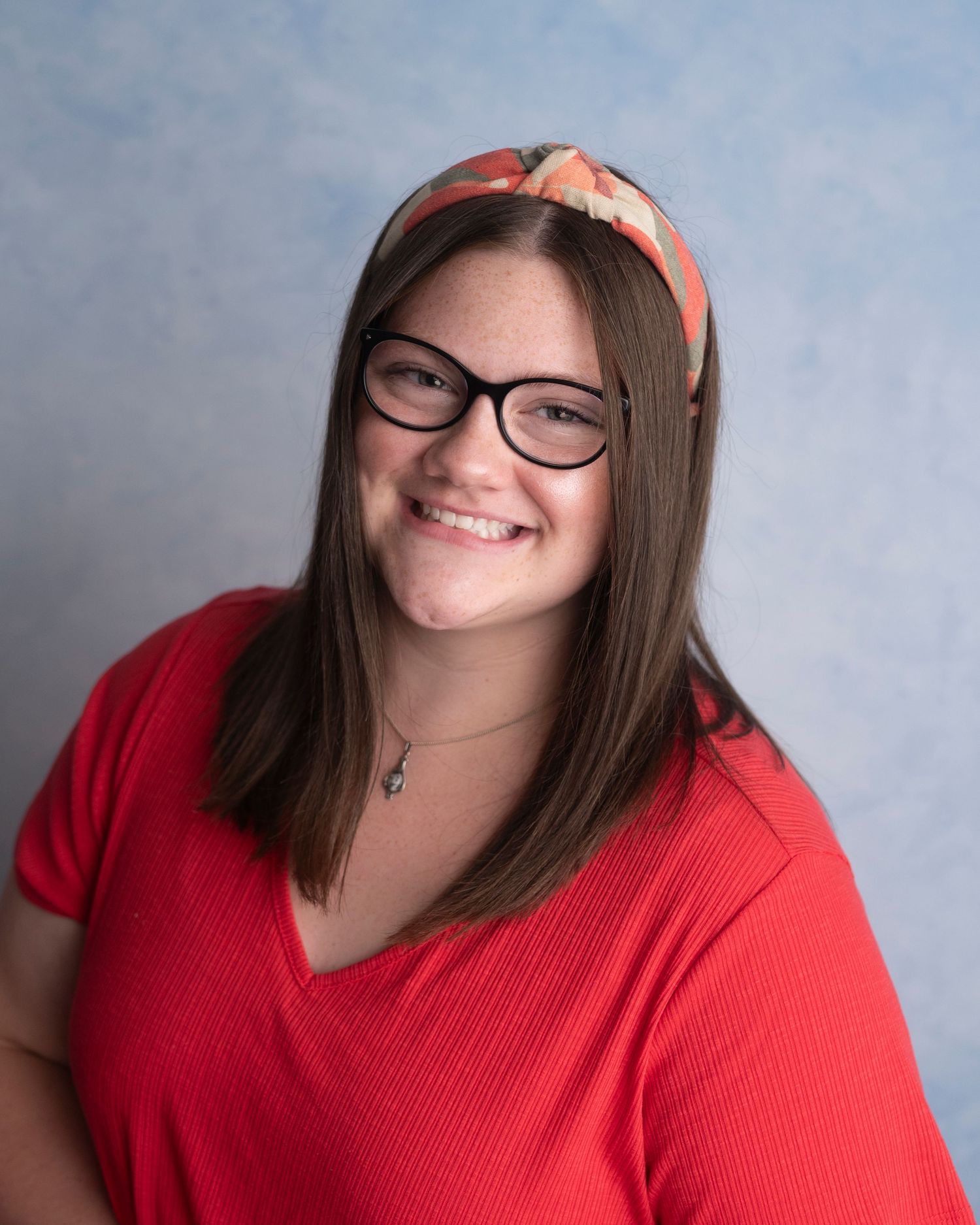 Portrait of someone wearing glasses and a peach headband with long brown hair smiling in a red v-neck top.