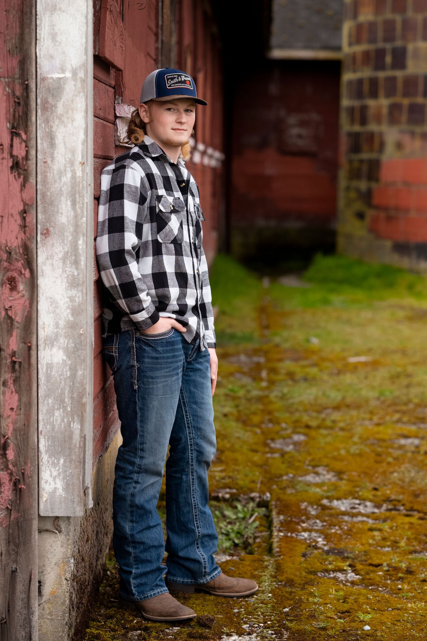 Young boy in buffalo plaid shirt and jeans poses against rustic brick wall in autumn outdoor setting.